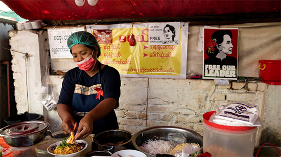 امرأة تطبخ في كشك للطعام عليه ملصقات ضد الانقلاب العسكري في يانغون